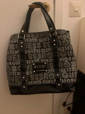 Svart handväska från Guess - Snygg svart handväska från Guess med tydligt logomönster i vitt över hela väskan. Väskan har dubbla handtag i konstläder, silverfärgade detaljer och en Guess-plakett framtill. Materialet är en mix av textil och konstläder. Perfekt för dig som gillar statement-accessoarer.