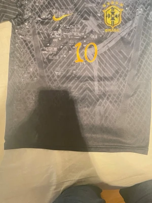 Svart Brasilien fotbollströja Nike - Snygg svart Brasilien fotbollströja från Nike med gult tryck, nummer 10 på bröstet och CBF-emblem. Tröjan har ett unikt mönster i tyget och är tillverkad i ett lätt och ventilerande material, perfekt för fotbollsträning eller match.