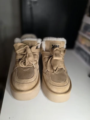 Beiga stövletter med fuskpäls från Xti - Snygga beiga stövletter från Xti med grov sula, breda snören och mysig vit fuskpäls runt kanten. Ovandelen är i mockaliknande material och textil, med detaljer i metall på sidan. Kan vara svåra att gå i för vissa pga hård sula. Använda ca 3 gånger.