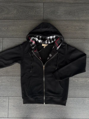 Svart hoodie från Burberry med dragkedja - Svart hoodie från Burberry med hel dragkedja och klassiskt rutigt foder i huvan. Hoodien har fickor framtill och justerbar dragsko i huvan. Perfekt för en stilren och trendig look.