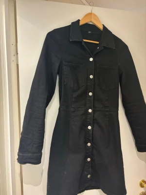 Svart jeansklänning med knappar - Svart kort denim klänning  med långa ärmar och silverfärgade knappar hela vägen framtill. Klänningen har två bröstfickor och klassisk krage, perfekt för en cool och avslappnad stil.