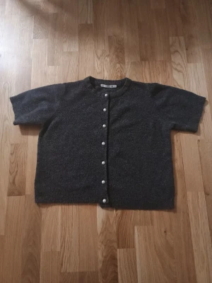 Mörkgrå kofta med pärlknappar - Kortärmad mörkgrå kofta med rund halsringning och pärlknappar framtill. Koftan har en rak passform och ribbstickade kanter vid ärmslut och nederkant. Perfekt för lager-på-lager och enkel att styla till olika looks.