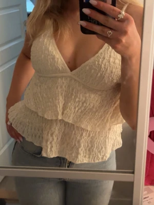 Krämvit linnetopp med volanger - Säljer en krämvit linnetopp med volangdetaljer och halterneck. Toppen har en lätt strukturerad yta och djup v-ringning. Perfekt för varma dagar och ger en luftig känsla.