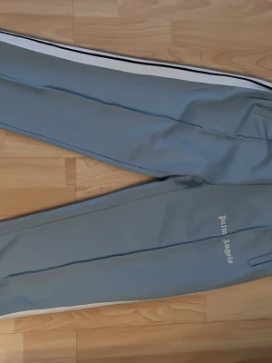 Blåa mjukisbyxor Palm Angels - Snygga ljusblå mjukisbyxor från Palm Angels med vita sidoränder och logga tryckt på benet. Byxorna har raka ben, resår i midjan och dragkedjor vid bensluten. Perfekta för en chill och sportig look. Storlek L men passar även för M.