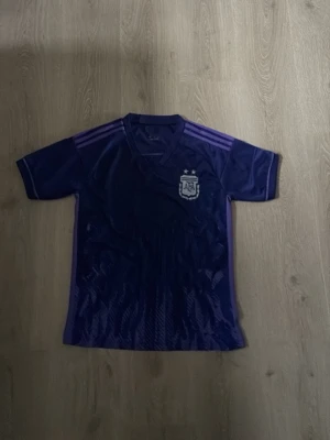 Lila fotbollströja Argentina Adidas - Snygg lila fotbollströja från Adidas med Argentinas landslagsmärke på bröstet. Tröjan har korta ärmar, V-ringning och tre klassiska Adidas-ränder på axlarna i en ljusare lila nyans. Perfekt för träning eller match.