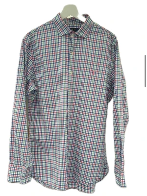 Rutig skjorta från Polo Ralph Lauren - Klassisk långärmad skjorta från Polo Ralph Lauren i röd, blå och grön rutig bomull. Skjortan har knappar framtill och en liten broderad logga på bröstet. Perfekt för en avslappnad men ändå stilren look.
