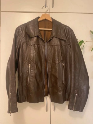 Brun skinnjacka med dragkedjor - Snygg brun skinnjacka med klassisk krage och dragkedjor framtill samt på ärmarna. Jackan har två fickor med dragkedja och dekorativa spännen baktill för extra stil. Perfekt för dig som vill ha en tidlös och cool look. Min mosters gammla så skicket kanske inte är det bästa fodret på insidan börjar lossna men syns inte mycket alls! Det stod ej nån storlek men skulle säga M/38