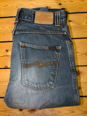 Blå jeans från Nudie Jeans - Säljer ett par klassiska blå jeans från Nudie Jeans med snygga slitningar och kontrastsömmar. Jeansen har raka ben, fem fickor och en ikonisk broderad bakficka. Tillverkade i slitstark denim med cool tvättad look.
