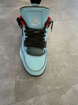 Blåa Air Jordan 4 sneakers - Snygga Air Jordan 4 sneakers i ljusblå mock per med svarta detaljer och röda insidor. Skon har svart nät på tån och sidan, samt vita och svarta sulor. Ikonisk Jumpman-logga i guld på plösen. Perfekt för dig som vill sticka ut med stilrena och sportiga sneakers.