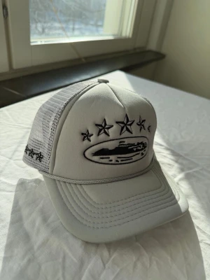 Grå meshkeps med broderade stjärnor - Säljer en stilren grå keps med mesh på sidorna och bak, samt broderade svarta stjärnor och ett ovalt motiv framtill. Kepsen har böjd skärm och justerbar snapback. Perfekt för dig som vill ha en unik och trendig accessoar.