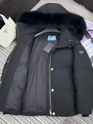 Svart dunjacka med pälskant från Prada - Snygg svart dunjacka från Prada med stor huva och lyxig svart pälskant. Jackan har quiltad design, dragkedja och tryckknappar framtill samt Pradas ikoniska trekantslogga på ärmen. Perfekt för kalla vinterdagar och riktigt trendig look.