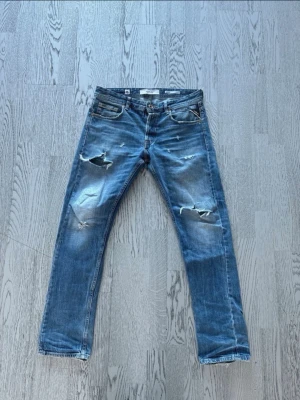 Blå slitna jeans från Replay - Säljer ett par blå jeans från Replay med slitningar och hål på låren och knäna. Jeansen har fem fickor, normal midja och raka ben. Materialet är klassisk denim i bomull. Perfekta för en avslappnad och trendig look.