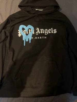 Palm Angels svart hoodie med blått hjärta - Svart hoodie från Palm Angels med stor vit logga och texten 'ST. BARTH' på bröstet. Ett stort blått hjärta med drip-effekt är tryckt över loggan. Klassisk huva och magficka framtill. Perfekt för dig som gillar streetwear och statement-plagg.