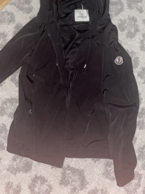 Svart vindjacka från Moncler - Snygg svart vindjacka från Moncler med huva och dragkedja framtill. Jackan har Moncler-logga på ärmen och en randig detalj på huvan. Tillverkad i lätt och tunt material, perfekt för blåsiga dagar. Två fickor med dragkedja och justerbar huva.