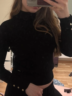 Svart polotopp med guldknappar - Snygg svart polotopp med långa ärmar och dekorativa guldknappar vid ärmsluten. Toppen har en tight passform och är perfekt att styla med höga jeans eller kjol. Enkel och stilren design som passar till många looks.