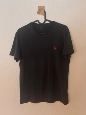 Svart t-shirt från Polo Ralph Lauren - Klassisk svart t-shirt från Polo Ralph Lauren med röd broderad logga på bröstet. T-shirten har rund halsringning och korta ärmar. Tillverkad i mjuk bomull för en skön känsla och enkel stil.