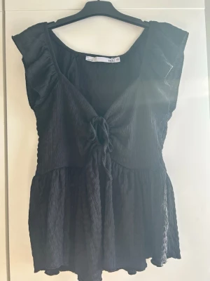 Svart volangtop med knyt från 157 - Snygg svart topp från 157 med volangdetaljer vid axlarna och v-ringning. Toppen har knyt framtill och är lätt rynkad i materialet för en luftig känsla. Perfekt till jeans eller kjol för en trendig look.