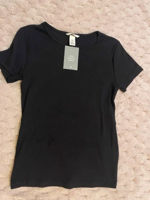 Svart ribbad t-shirt från H&M - En svart, ribbad t-shirt från H&M i storlek L. Tillverkad av 95% ekologisk bomull och har en klassisk rund halsringning samt korta ärmar. Perfekt basplagg som är både enkel och stilren. Strl L men funkar på mindre beroende på vad man vill ha för passform 