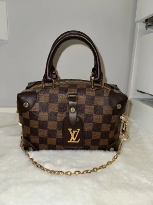 brun damier handväska - Snygg handväska i klassiskt brunt damier-mönster. Väskan har guldfärgade metalldetaljer, dubbla handtag och en avtagbar kedjerem. 