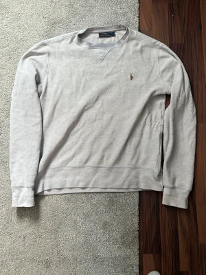 Ljusgrå sweatshirt från Polo Ralph Lauren - Klassisk ljusgrå sweatshirt från Polo Ralph Lauren med broderad logga på bröstet. Tröjan har rund halsringning, ribbade muddar och är tillverkad i mjukt bomullsmaterial. Perfekt för en avslappnad och stilren look.