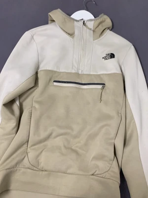 Beige hoodie från The North Face - Snygg beige hoodie från The North Face med halv dragkedja framtill och stor magficka med dragkedja. Hoodien har en huva och är tillverkad i ett mjukt material som känns skönt mot huden. Perfekt för en avslappnad streetwear-look.