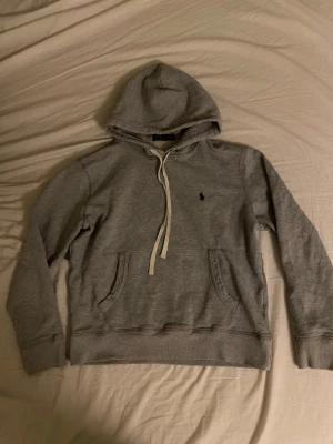 Grå hoodie från Polo Ralph Lauren - Snygg grå hoodie från Polo Ralph Lauren med klassisk huva och vita dragsnören. Har känguruficka framtill och den ikoniska lilla broderade loggan på bröstet. Perfekt för en chill och avslappnad stil. Mjuk bomullsblandning som är skön att ha på sig.