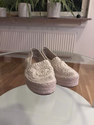 Beigea loafers/sommarskor med brodyr - Säljer ett par beige loafers/sommarskor i flätad design och snyggt broderat mönster på ovandelen. Skorna har tillverkade i tyg med detaljer som ger en bohemisk känsla. Perfekta för dig som gillar en avslappnad men trendig stil. Super att ha nu i vår/sommar. Använd endast 1 gång