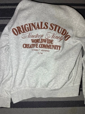 Grå hoodie från Originals Studio - Snygg ljusgrå hoodie från Originals Studio med stor brun text på ryggen och mindre logga på bröstet. Hoodien har huva och en klassisk magficka framtill. Tillverkad i mjukt sweatshirtmaterial, perfekt för en avslappnad och trendig look.