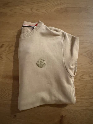 Beige sweatshirt från Moncler - Snygg beige sweatshirt från Moncler med rund hals och ribbade muddar. Tröjan har Moncler-logga broderad på bröstet och klassisk passform. Perfekt för dig som gillar stilrena och exklusiva plagg.