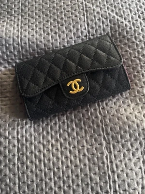 Svart quiltad handväska  - Snygg svart quiltad handväska  med klassiskt guldigt CC-spänne framtill. Väskan är tillverkad i läder med struktur och har magnetlås. Insidan har guldfärgad logga och detaljer i metall. Perfekt för dig som gillar lyxiga accessoarer.