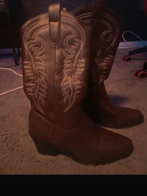 Bruna cowboy boots med broderi - Snygga bruna cowboy boots med dekorativt broderi längs skaftet. Klassisk spetsig tå och låg klack. Tillverkade i konstläder med dragflikar upptill för enkel påtagning. Perfekta för dig som vill ha westernstil i garderoben.