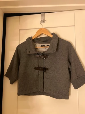 Grå trekvartsärmad kofta  - Snygg grå kofta från något märke som jag köpte på sellpy i french terry-material. Kortärmad och trekvarts ärmad modell med coola spännen framtill och hög krage. Perfekt för lager-på-lager och en chill streetstyle-look. Den är as snygg