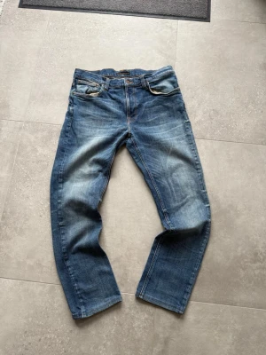 Blå jeans från Nudie Jeans - Säljer ett par klassiska blå jeans från Nudie Jeans med raka ben och snygga slitningar. Jeansen har fem fickor, orange sömmar och en patch bak i midjan. Tillverkade i denim av bomull och har en avslappnad passform. Modell lean dean=mer slim storlek 33w 32L. 