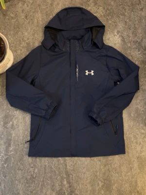 Under Armour - Snygg marinblå vindjacka från Under Armour med huva och dragkedja framtill. Jackan har två sidofickor med dragkedja. Helt ny. PRIS KAN DISKUTERAS 