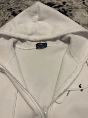 Vit hoodie från Polo Ralph Lauren - Klassisk vit hoodie från Polo Ralph Lauren med dragkedja framtill, justerbar huva med snören och två fickor. Liten broderad logga i marinblått på bröstet. Tillverkad i mjukt och bekvämt material, perfekt för en avslappnad stil.