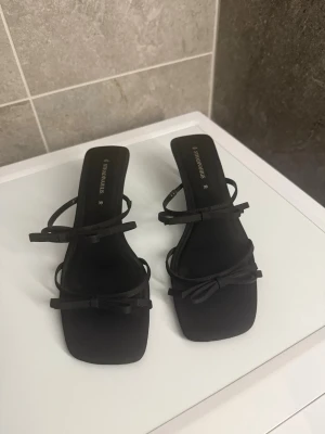 Svarta sandaler med klack från Stradivarius - Säljer ett par stilrena svarta klackar från Stradivarius med fina rosettdetaljer framtill. Låg klack. Storlek 39. Passar fint. Snygga till fest. 💋