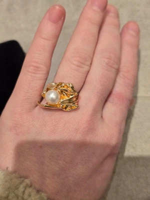 Guldfärgad ring med pärla - Unik guldfärgad ring med en stor vit pärla i mitten. Ringen har en organisk, vågig form som ger ett coolt och trendigt intryck. Perfekt för dig som gillar statement-smycken och vill sticka ut. Materialet är metall och pärla. Cirka storlek 16.5