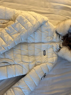 Vit pufferjacka med fuskpäls från Marikoo - Snygg vit pufferjacka från Marikoo med avtagbar huva och fuskpälsdetalj. Jackan har quiltad design, dragkedja framtill och ribbade muddar vid ärmslut. Perfekt för kalla vinterdagar och riktigt trendig look. Storlek M