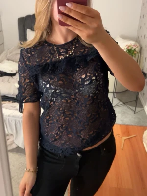 Mörkblå spetsblus med volang - Snygg mörkblå blus i spets med korta ärmar och volangdetalj över bröstet. Blusen är genombruten och har ett feminint mönster. Perfekt att styla med ett linne under eller som den är för en trendig look.