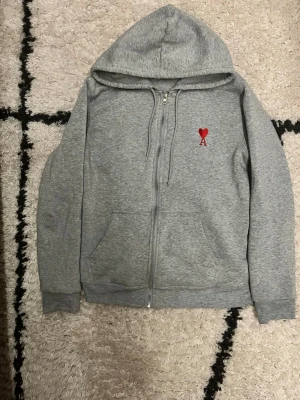 Grå hoodie med röd Ami - Snygg grå zip-hoodie från AMI Paris med rött Ami-de-Coeur broderat på bröstet. Klassisk modell med dragkedja, två fickor fram och justerbar huva. Tillverkad i mjuk bomullsblandning som är skön att ha på sig. Perfekt för en chill och stilren look.