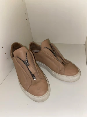 Beige arigatos - Snygga beige sneakers i läder med unik dragkedja framtill istället för klassiska snören. Skorna har en chunky vit sula och minimalistisk design som passar till många olika stilar. Perfekta för dig som gillar stilrena och moderna detaljer.