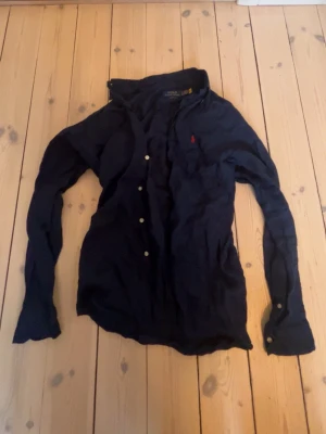 Mörkblå linneskjorta från Ralph Lauren - Klassisk mörkblå linneskjorta skjorta från Ralph Lauren med lång ärm och knappar framtill. Skjortan har den ikoniska röda logotypen broderad på bröstet. Perfekt för en stilren och avslappnad look. Väldigt skön på sommaren eller varmt väder! Måste strykas men annars i väldigt bra skick!