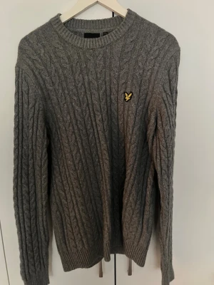 Grå stickad tröja från Lyle & Scott - Stickad grå tröja från Lyle & Scott präglad med deras klassiska gula örnlogga på bröstet. Tröjan har rund halsringning, långa ärmar och ribbade muddar vid ärmslut och nederkant. Perfekt för lager på lager.