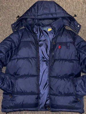 Marinblå pufferjacka Polo Ralph Lauren - Snygg marinblå pufferjacka från Polo Ralph Lauren med avtagbar huva och dragkedja framtill. Jackan har två sidofickor med dragkedja och den klassiska röda loggan broderad på bröstet. Perfekt för kalla vinterdagar och riktigt stilren.