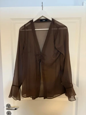 Brun transparent blus med volangärm - Snygg brun transparent blus från Bik Bok med långa ärmar och volangdetaljer vid ärmslut. Blusen har en djup V-ringning och knytning framtill, vilket ger en trendig och luftig look. Perfekt att styla lager på lager.