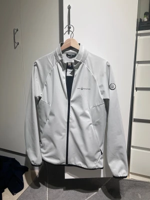 Sail racing jacka - Snygg ljusgrå vindjacka från Sail Racing med svart dragkedja och diskret logga på bröstet. Jackan har hög krage, två sidofickor med dragkedja och ett emblem på ärmen. Tillverkad i ett lätt och funktionellt material som passar perfekt för blåsiga dagar. De finns nån flöck hör och där som förmodligen går bort i tvätt. Pris går självklart att diskutera