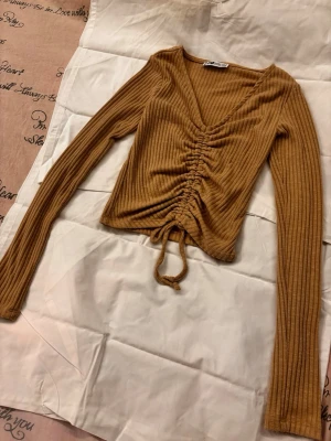 Ribbad beige/brun croppad topp med snörning - Snygg ribbad topp i brun beige/färg med croppad passform och långa ärmar. Toppen har v-ringning och snörning framtill som ger en rynkad effekt. Perfekt för dig som gillar trendiga detaljer och en bekväm stil.