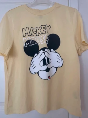 Gul Mickey Mouse t-shirt - Snygg gul t-shirt med tryck av Mickey Mouse på framsidan där han håller händerna för ögonen. T-shirten har rund hals och korta ärmar. Perfekt för dig som gillar Disney och vill ha en lekfull stil.