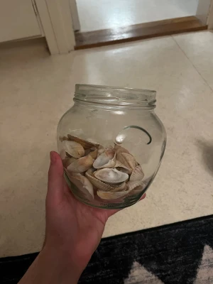 Glasburk med snäckskal - Säljer en rund glasburk fylld med olika snäckskal i beige, vitt och ljusbrunt. Perfekt som dekoration eller pysselmaterial. Burken är genomskinlig och har en bred öppning som gör det enkelt att ta ut snäckskalen.