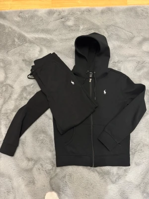 Polo Ralph Lauren set - Säljer ett svart loungewear-set från Polo Ralph Lauren med zip-hoodie och matchande byxor. Setet har den klassiska vita loggan broderad på både tröjan och byxan. Hoodien har dragsko och fickor, materialet är mjukt och skönt, perfekt för chill dagar hemma.